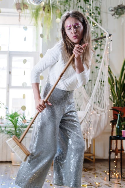 A person is standing on a wooden floor, surrounded by scattered gold confetti, with bare feet visible. They are holding a broom and standing near a dustpan and brush set, preparing to collect the conf