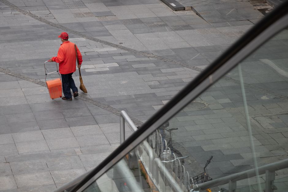 A street-level view of a pedestrian sidewalk in Putney, showing a person dressed in an orange jacket, blue trousers, and a red cap sweeping the pavement with a broom while carrying an orange waste collection bin. The surface consists of large, rectangular grey paving stones with some darker patches, indicating recent cleaning or wear. Visible elements include a metal railing, partially obscured bicycles parked alongside, and a reflective glass storefront or window in the foreground, adding depth to the scene. Natural daylight illuminates the area, highlighting the clean, smooth surface of the stones and the overall tidiness of the environment, illustrating a typical urban street scene in need of routine maintenance. This image aligns with the services offered by Putney Cleaner for surface cleaning and deep cleaning on commercial shopfronts, as featured in the case study on gum removal on Putney High Street.