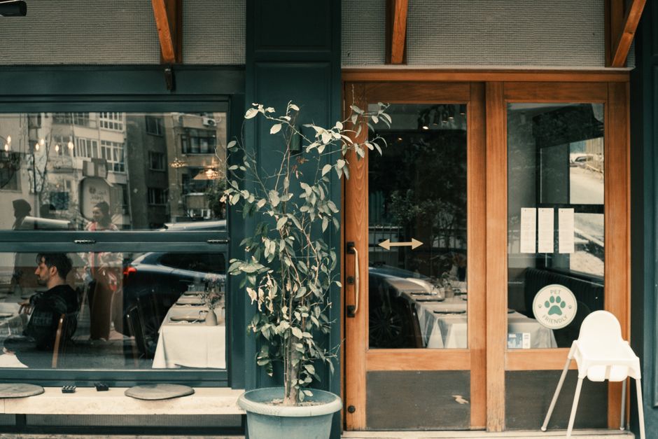 The image depicts the exterior of a commercial shopfront with large glass windows and a wooden door. The glass surfaces are clean and transparent, reflecting the street scene behind, including parked cars and pedestrians. Outside, there is a potted plant with green and silver leaves placed on the pavement, adding a decorative touch to the storefront. The window sill appears tidy, with no visible dust or dirt. The interior of the shop, visible through the glass, shows neatly arranged tables covered with white tablecloths, indicating a restaurant or café setting. The lighting inside is bright, emphasizing the cleanliness of the space. The overall appearance suggests recent or ongoing surface cleaning and maintenance, consistent with the professional cleaning services provided by Putney Cleaner. This image highlights the importance of surface cleaning, hygiene, and presentation in commercial premises, demonstrating a well-maintained shopfront.