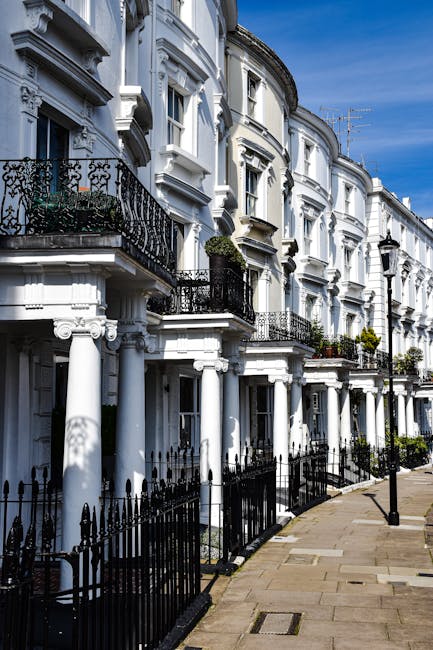 View of a row of historic white Victorian terraced houses on Lower Richmond Road in Putney, featuring ornate architectural detailing, black wrought iron balconies and railings, and large sash windows. The pavement running alongside is clean and well-maintained, with a black metal fence separating the street from the front gardens. Bright daylight illuminates the facades, highlighting the pristine condition of the building exteriors. This image reflects a well-preserved residential street, emphasizing the importance of surface cleaning and maintenance for optimal appearance. For professional cleaning services in Putney, contact Putney Cleaner for expertise in domestic cleaning, deep cleaning, and surface sanitisation.