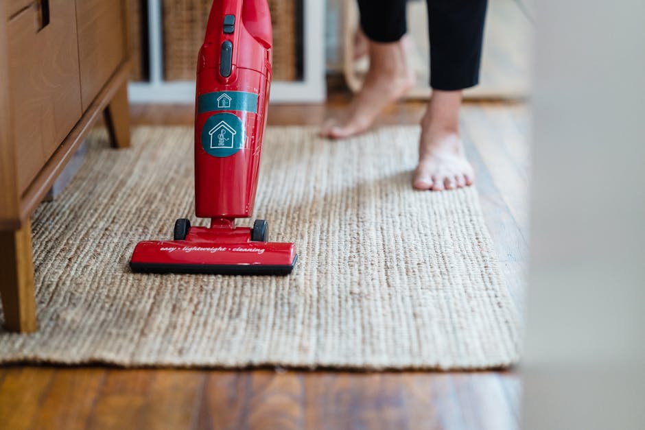 Close-up of a red handheld vacuum cleaner placed on a beige textured carpet in a domestic setting, with a person's bare feet and black leggings visible in the background. The carpet appears clean and well-maintained, situated near wooden flooring and a piece of wooden furniture. The lighting is natural, highlighting the carpet's fibers and the shiny surface of the vacuum. This image illustrates surface cleaning and deep cleaning practices, emphasizing hygienic upkeep of residential carpets. The logo and branding on the vacuum suggest professional cleaning services, and the scene is associated with domestic cleaning and sanitisation, as promoted by Putney Cleaner near Putney Pier. The focus is on the cleanliness and maintenance of the carpet, reflecting effective hygiene routines.