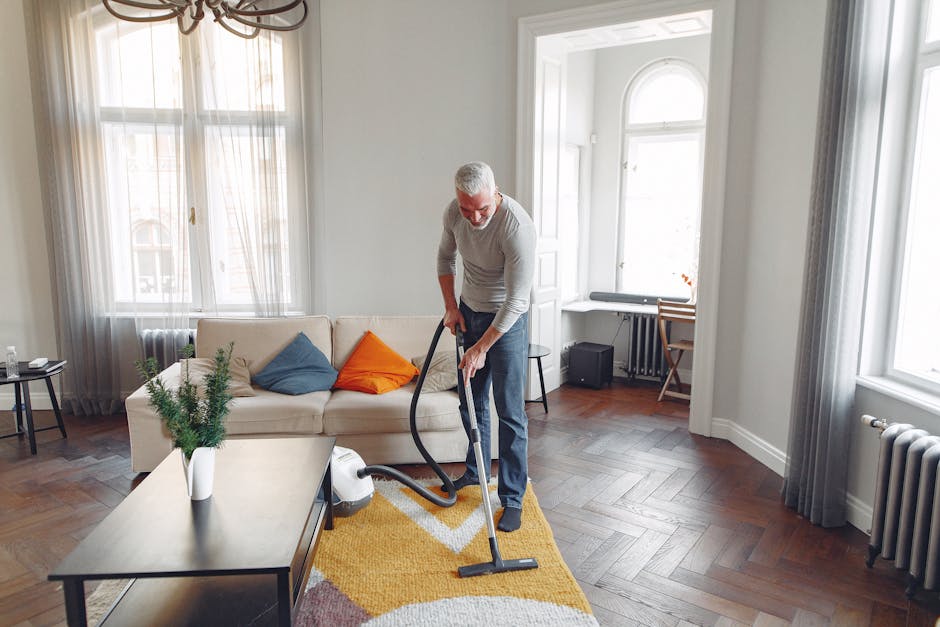 A middle-aged man with short gray hair and glasses is engaged in surface cleaning within a bright, modern living room. He is wearing a light gray long-sleeve shirt and dark pants, using a vacuum cleaner with a black hose attachment to clean a colorful, patterned rug on a wooden floor. The room features a beige sofa adorned with three cushions in blue, orange, and gray, placed against a large window with sheer white curtains that allow natural light to fill the space. To the side, there is a low black side table with a glass bottle and a small plant in a white pot. The walls are painted white, and the area is well-lit with ambient daylight, creating a clean and tidy atmosphere. Nearby, a small desk with a wooden chair and a radiator under the window are visible, emphasizing the room’s comfortable and orderly condition. The image highlights professional domestic cleaning processes, with attention to maintaining hygiene through thorough surface and carpet cleaning, demonstrating [COMPANY_NAME]’s expertise in deep cleaning and surface sanitisation.