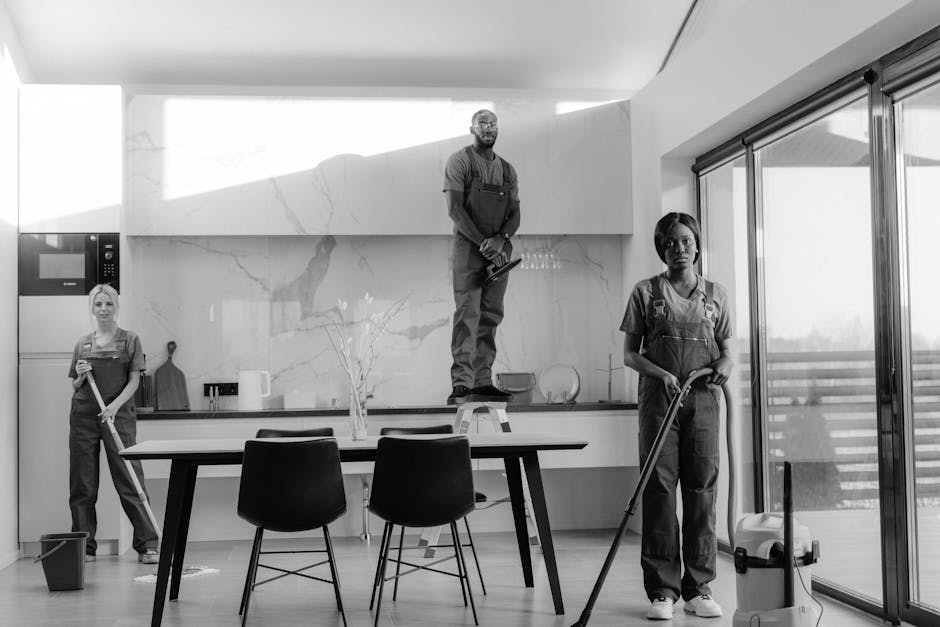 Two professional cleaners in a modern living room actively engaged in surface cleaning and deep cleaning tasks. The female cleaner on the left uses a vacuum cleaner with a hose attachment to clean the