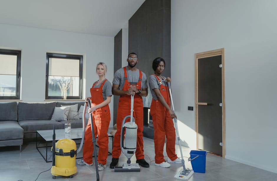 Three professional cleaners from Putney Cleaner are performing surface cleaning and sanitisation in a modern living room. They are dressed in grey t-shirts and orange overalls, with tools including a vacuum cleaner, mop, and bucket visible. The room features a grey fabric sofa with cushions, two black-framed windows allowing natural light, and a dark accent wall contrasting with light-colored walls. The floor appears to be made of large grey tiles, which are being cleaned with an upright vacuum and mop, highlighting a focus on deep and hygienic domestic cleaning. The scene emphasizes thorough cleaning processes and the importance of maintaining hygiene with well-equipped staff in a contemporary residential setting, aligning with safety and licensing considerations discussed on the 'Do private cleaners need licences in Putney?' page. Putney Cleaner’s professional team is dedicated to delivering comprehensive surface cleaning and sanitisation, ensuring all surfaces are left clean, shiny, and dust-free.