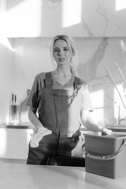A young woman in casual attire and work overalls is seen standing in a modern kitchen with a clean, shiny stone or marble backsplash. She is holding a white cloth in her right hand and resting her left hand on a plastic container on the kitchen countertop. The surface of the counter appears smooth and well-maintained, with a neat arrangement of utensils and tools visible in the background. Bright natural light illuminates the space, highlighting the pristine, dust-free surface and the reflection on the cooker hood and sink area. This scene illustrates domestic surface cleaning and deep cleaning practices, aligning with the themes of hygiene and maintenance promoted by Putney Cleaner. The overall setting emphasizes professional cleaning standards related to safety and sanitisation, suitable for domestic cleaning service promotion.