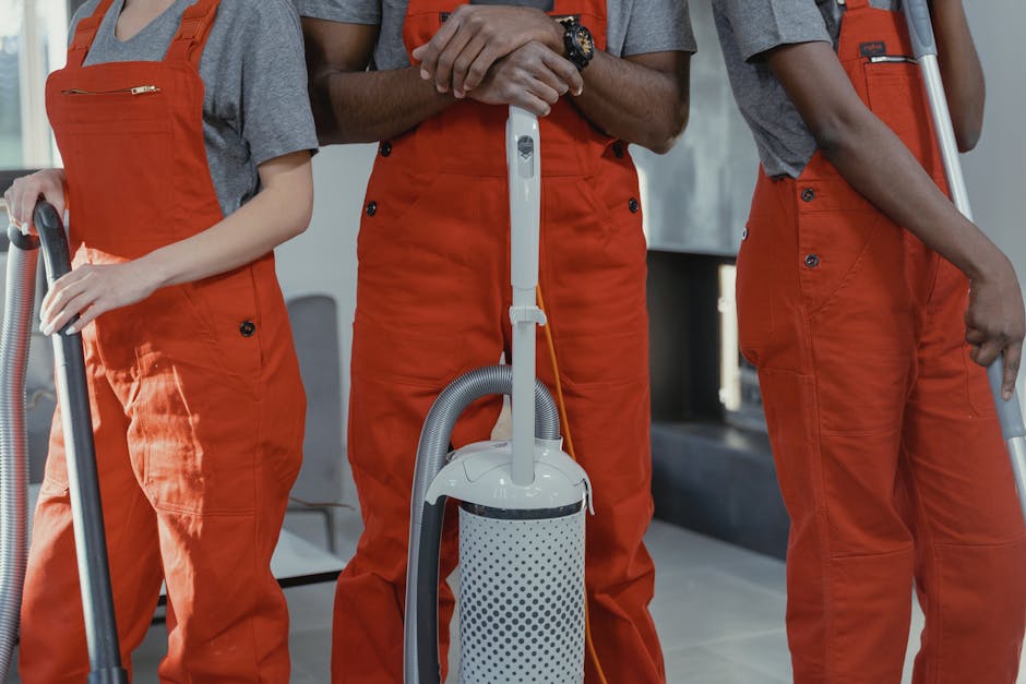Three professional cleaners from Putney Cleaner are performing surface cleaning and sanitisation in a modern living room. They are dressed in grey t-shirts and orange overalls, with tools including a vacuum cleaner, mop, and bucket visible. The room features a grey fabric sofa with cushions, two black-framed windows allowing natural light, and a dark accent wall contrasting with light-colored walls. The floor appears to be made of large grey tiles, which are being cleaned with an upright vacuum and mop, highlighting a focus on deep and hygienic domestic cleaning. The scene emphasizes thorough cleaning processes and the importance of maintaining hygiene with well-equipped staff in a contemporary residential setting, aligning with safety and licensing considerations discussed on the 'Do private cleaners need licences in Putney?' page. Putney Cleaner’s professional team is dedicated to delivering comprehensive surface cleaning and sanitisation, ensuring all surfaces are left clean, shiny, and dust-free.