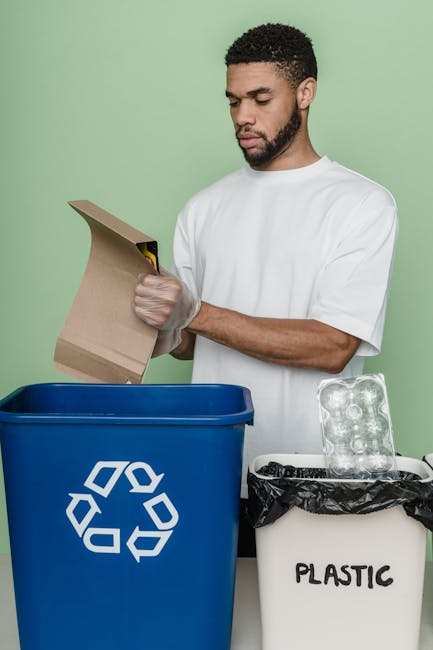 A man with a short curly hairstyle and a beard, dressed in a white t-shirt, is seen disposing of a cardboard box into a large blue recycling bin with a white recycling symbol, designated for plastic recycling. He is standing upright with both hands holding the cardboard box, positioned in front of a light green background, suggesting a clean indoor environment. To his right, there is a smaller white bin marked 'PLASTIC' with a black garbage bag lining it, containing empty plastic bottles, and a partially visible black item, possibly a trash bag or cleaning cloth. The scene emphasizes proper waste separation and recycling practices in a domestic setting, and the cleaning company Putney Cleaner's focus on environmentally friendly disposal methods aligns with eco-friendly practices related to mattress disposal and recycling. The surface appears clean and well-organized, with bright, even lighting enhancing visibility of the recycling process.