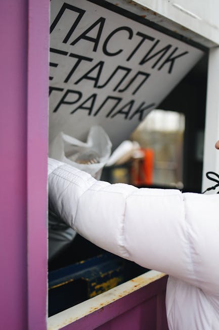 A person reaching into a recycling bin under a sign with multilingual text, possibly in a public or commercial setting. The bin is purple, and the person's arm is covered with a white sleeve, indicating they are wearing a protective or uniform garment. The background shows a black surface with some visible items like a white plastic bag and an orange object, suggesting a waste disposal or recycling station. The lighting is natural, and the surfaces, including the purple bin, appear clean and well-maintained. The sign above the bin features bold black text in multiple languages, emphasizing waste separation, consistent with eco-friendly initiatives like mattress disposal and recycling promoted by Putney Cleaner. This image relates to maintaining hygiene and proper waste management in accordance with professional cleaning standards, as seen on the page about eco-friendly mattress disposal and recycling in Putney.