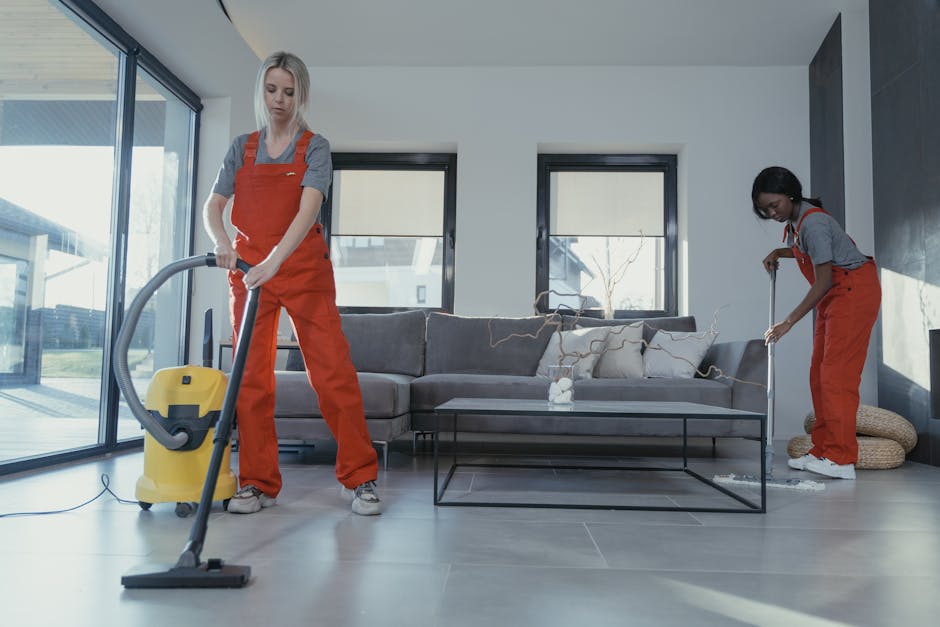 Two professional cleaners wearing orange overalls are performing surface cleaning in a modern living room. The cleaner on the left is vacuuming a large, light gray tiled floor using a yellow vacuum cl