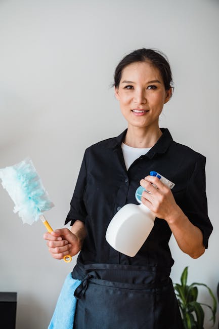 Two professional cleaners wearing orange overalls are performing surface cleaning in a modern living room. The cleaner on the left is vacuuming a large, light gray tiled floor using a yellow vacuum cl