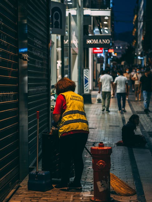 A sanitation worker wearing a yellow high-visibility vest marked 'Dublin Administration' is performing surface cleaning on a dark metal shutter of a commercial building on a busy urban street at night. The worker is using a cloth or spray bottle to wipe the shutter, which is closed, with a small black trolley or cleaning cart nearby. The tiled sidewalk is clean, with some street furniture, including a red fire hydrant and a trash bag, visible in the foreground. In the background, illuminated shop signs, including 'ROMANO', and pedestrian activity with people walking along the street are visible, suggesting a vibrant commercial area. The bright lighting highlights the shiny surfaces and contributes to a well-maintained and hygienic environment, aligning with deep cleaning and sanitisation principles promoted by Putney Cleaner in their office deep-clean checklist for Putney High Street businesses.