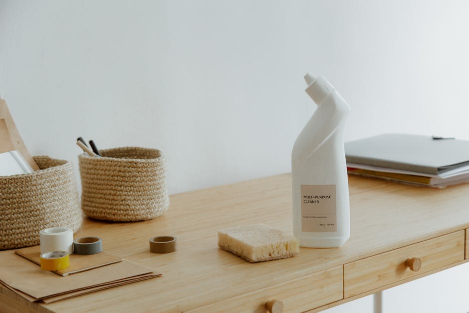 A wooden desk with a light natural finish is shown in a well-lit room. On the desk, there is a white multi-purpose cleaner spray bottle with a beige label, a natural-colored sponge, and a few open rolls of tape in white, gray, and yellow. Behind these items, two woven baskets are visible, containing pens and small objects. To the right, a stack of neatly arranged papers or documents rests on the desk, all positioned against a plain white wall. The surface appears clean and free of dust or stains, indicating recent or ongoing surface cleaning. The overall scene reflects a tidy workspace with an emphasis on hygiene and maintenance, consistent with professional domestic cleaning standards offered by Putney Cleaner.