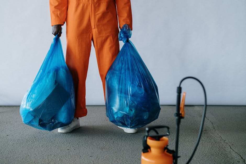 A person dressed in orange work overalls standing on a grey carpeted floor, holding two large blue plastic garbage bags filled with waste. The person's white shoes are visible at the bottom of the image. In the foreground, there is a black and orange handheld device connected to a cord, possibly a disinfectant spray or cleaning equipment. The background is plain with a light grey wall, emphasizing the scene of waste collection and disposal, consistent with surface cleaning and waste removal services as featured on putneycleaner.org.uk for the page about same-week rubbish removal quotes in Putney SW15 estates, provided by Putney Cleaner.