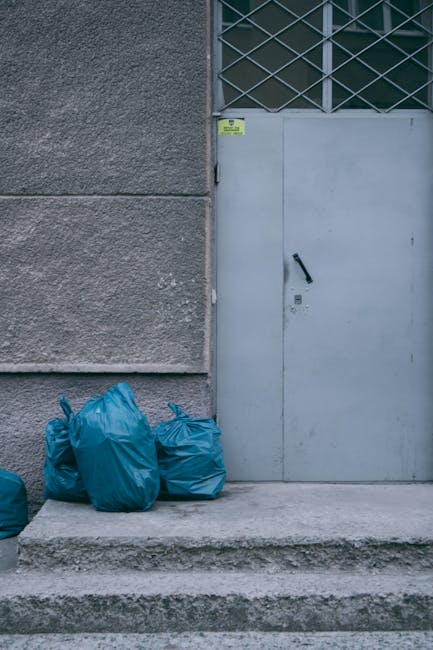 A person dressed in orange work overalls standing on a grey carpeted floor, holding two large blue plastic garbage bags filled with waste. The person's white shoes are visible at the bottom of the image. In the foreground, there is a black and orange handheld device connected to a cord, possibly a disinfectant spray or cleaning equipment. The background is plain with a light grey wall, emphasizing the scene of waste collection and disposal, consistent with surface cleaning and waste removal services as featured on putneycleaner.org.uk for the page about same-week rubbish removal quotes in Putney SW15 estates, provided by Putney Cleaner.